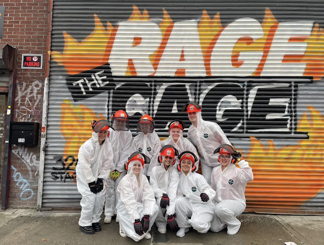 NYU Silver students wearing hard hats and white jumpsuits stand in front of a gratified garage door that says, “The Rage Cage.”